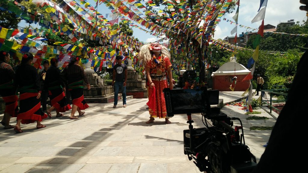 Camera operator filming a performer in a red costume under prayer flags at a heritage site in Nepal